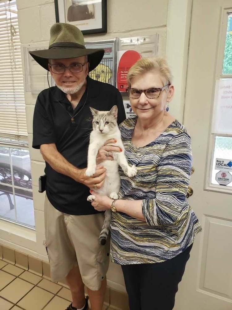 A couple holds a light-colored cat indoors. The man wears a hat; woman wears glasses.