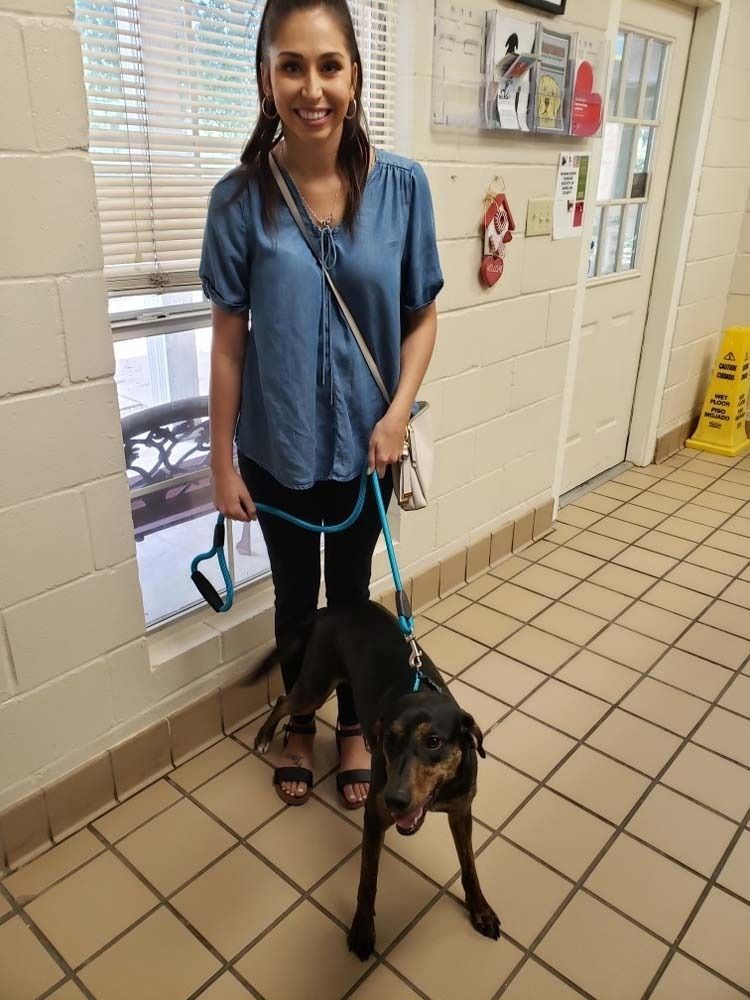 Woman with a dog on a leash in a tiled-floor hallway, smiling. The dog is black with brown markings.