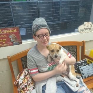 Woman in glasses and beanie holding a small dog on a bench; dog is brown and white.