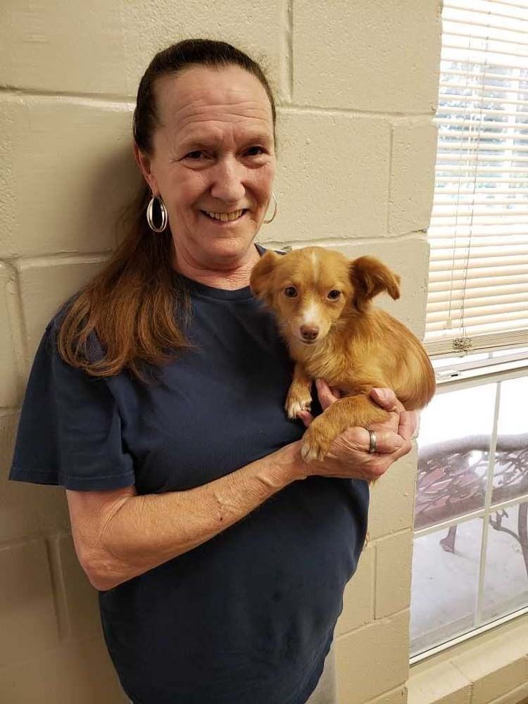 Woman in blue shirt holding small tan dog. Leaning against a beige wall, smiling.