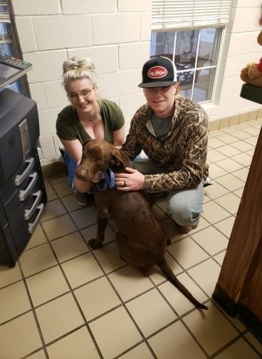 Two people kneeling with a brown dog in a room. The dog has a blue collar.
