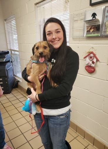 Woman holding a smiling, tan-colored dog. They are indoors with a window and a red leash.