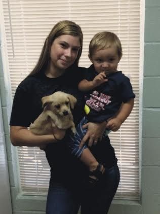Woman holding a small puppy and a child in front of a window with blinds.
