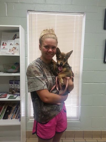Woman holding small brown dog, smiling. Background: window, shelves with brochures.