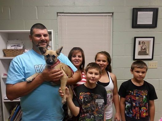 A man holds a puppy; four children pose beside them indoors with a window and shelves.