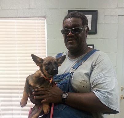 Man in overalls and sunglasses holding a puppy with a pink collar; indoor setting.