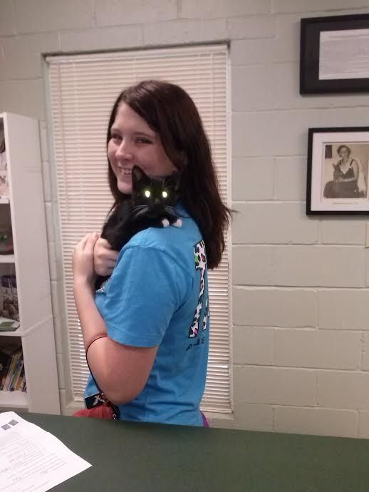 Woman in a blue shirt holding a black cat. The cat sits on her shoulder with glowing eyes.