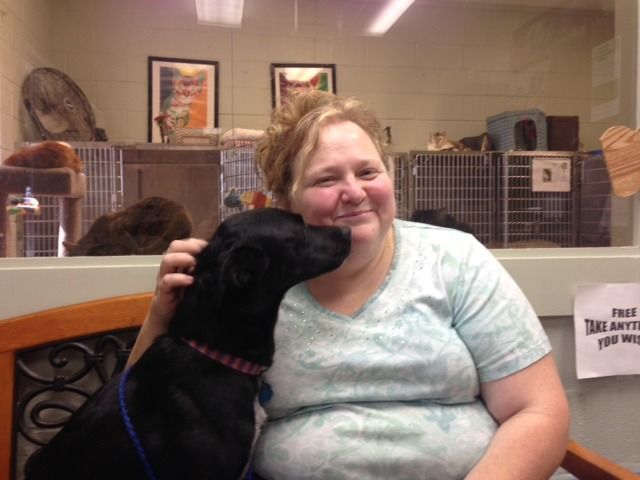 Woman smiles as black dog nuzzles her face inside a shelter. Other animals and cages visible in background.