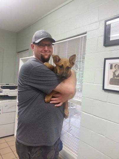 Man holding a brown puppy; they're inside near a window, wall, and copier. The man smiles.