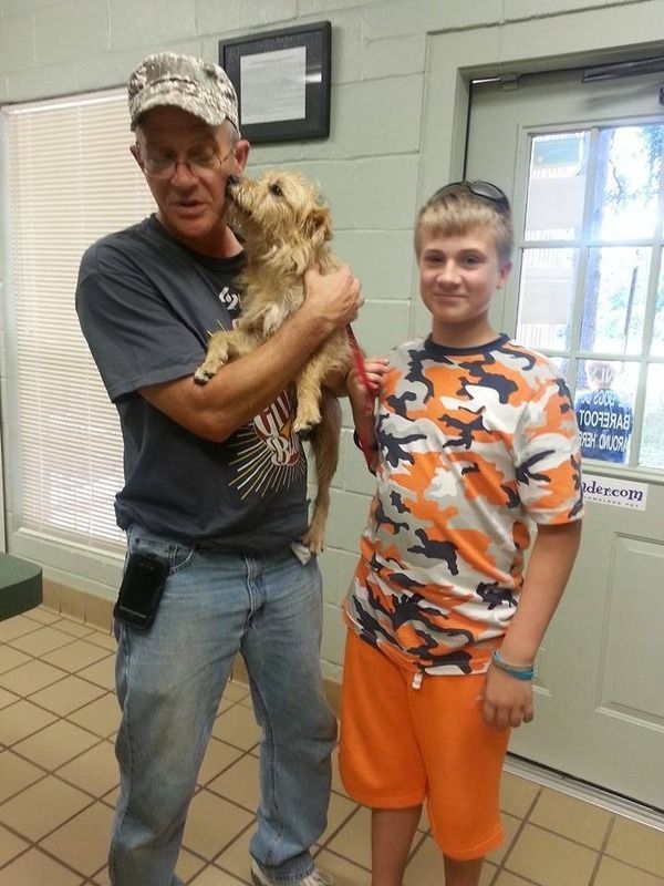 Man holding small dog, smiling, with boy in orange camo shirt standing next to him indoors.