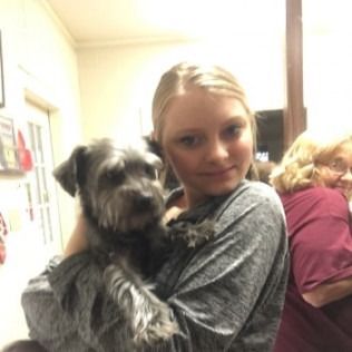 Young person holding a small, gray dog, smiling indoors. Another person in the background.