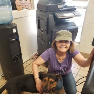Woman kneeling with a dog near a copier and water cooler. The woman wears a purple shirt and hat; the dog is black and brown.