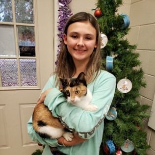 Girl holding a calico cat in front of a Christmas tree. Both are smiling.