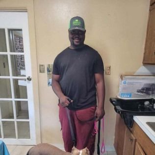 Man in black shirt and maroon pants stands in kitchen holding dog leashes, a dog at his feet.