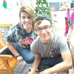 Two people smiling, posing together. Woman in tie-dye shirt sits on bench. Man in glasses and hat kneels. Holiday decorations in background.