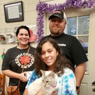 Family indoors with cat, woman in Red Hot Chili Peppers shirt, man in cap, girl holding cat, smiling.