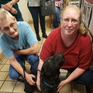 A dog kneels with two people, one on each side, in a hallway. The dog is black, and they are petting it.