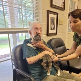 An older person sits with a cat in their lap, being assisted by a person beside them in a room.