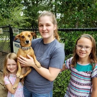 Woman holding a brown dog, with two girls smiling beside them near a fence.