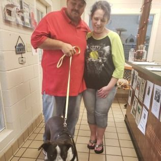 Couple adopting a dog at a shelter. Man holds leash, woman smiles. Interior, linoleum floor.