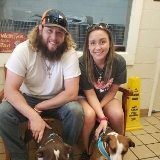 Couple sits with two dogs in an animal shelter; smiling, seated on a bench, looking at the camera.