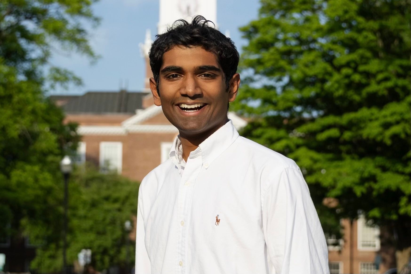 Man smiling, wearing a white shirt, standing outside in front of a brick building and green trees.