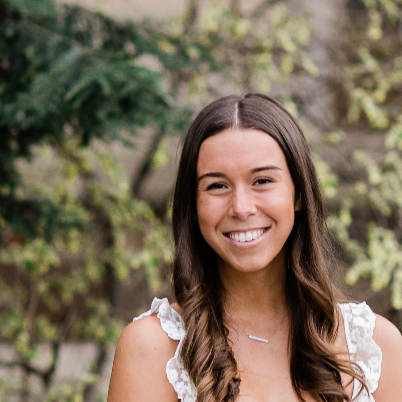 Woman smiling at the camera, wearing white lace top and silver necklace. Outdoors with greenery in background.