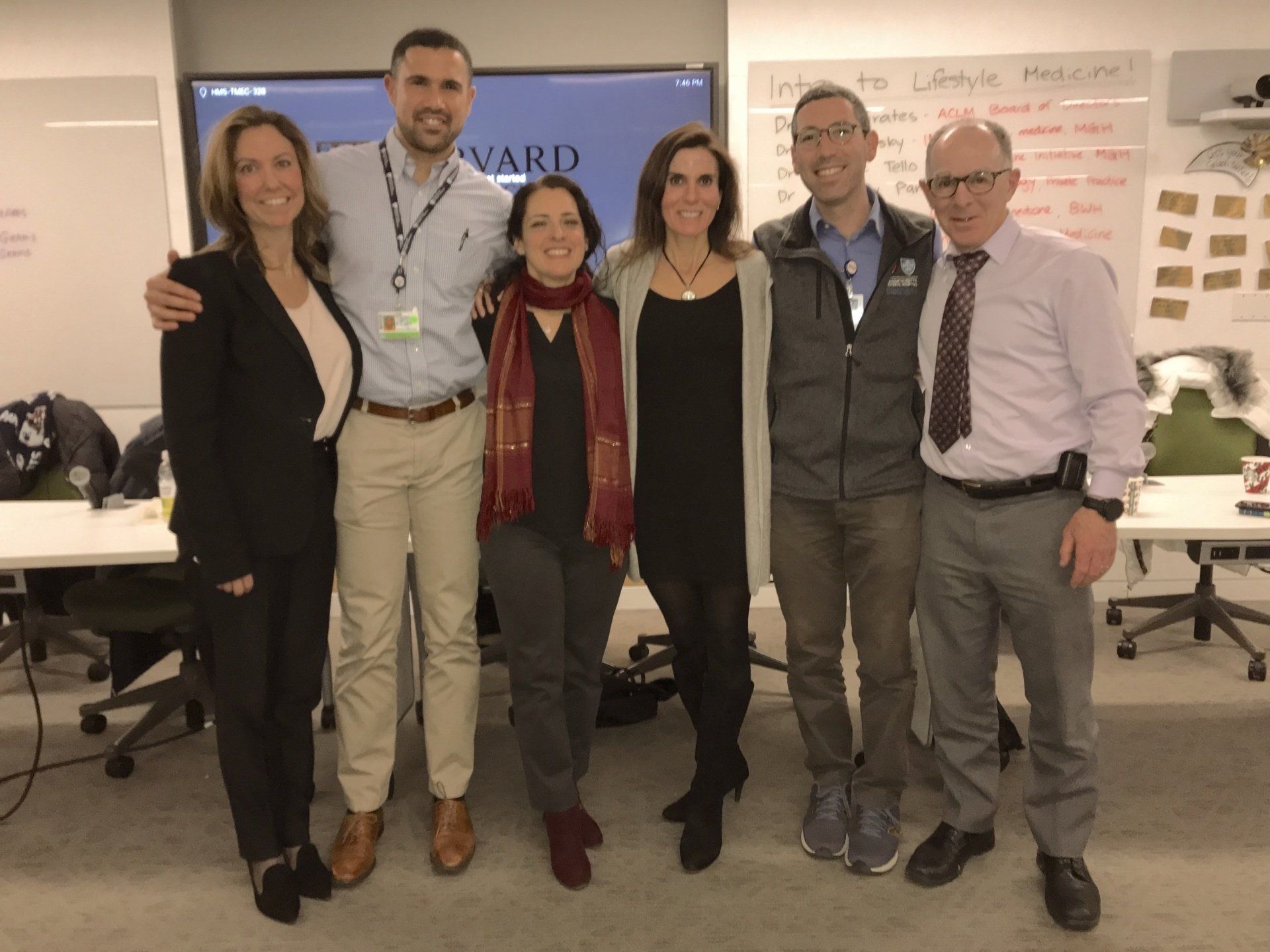 Six people standing together, smiling in an office setting. They are posing in front of a screen and whiteboard.