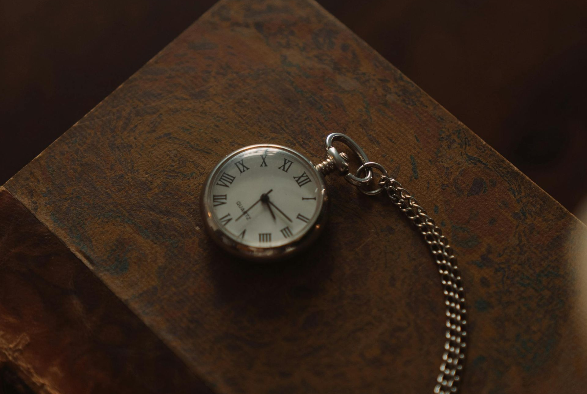 Pocket watch on aged book. Silver watch with white face, chain extending. Brown background.
