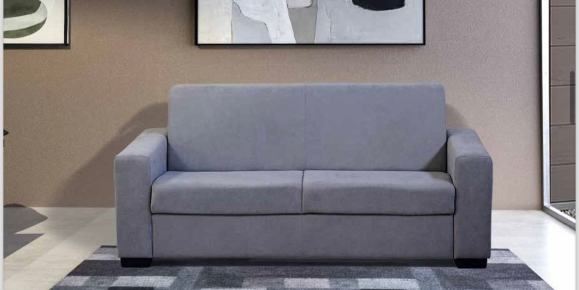 Un divano a due posti di colore grigio chiaro, dalle linee semplici e moderne, posizionato su un tappeto scuro a motivi geometrici in una stanza minimalista.