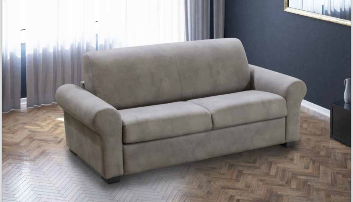 Un divano grigio chiaro a due cuscini con braccioli arrotondati, posizionato in una stanza con pavimento a spina di pesce e tende trasparenti.