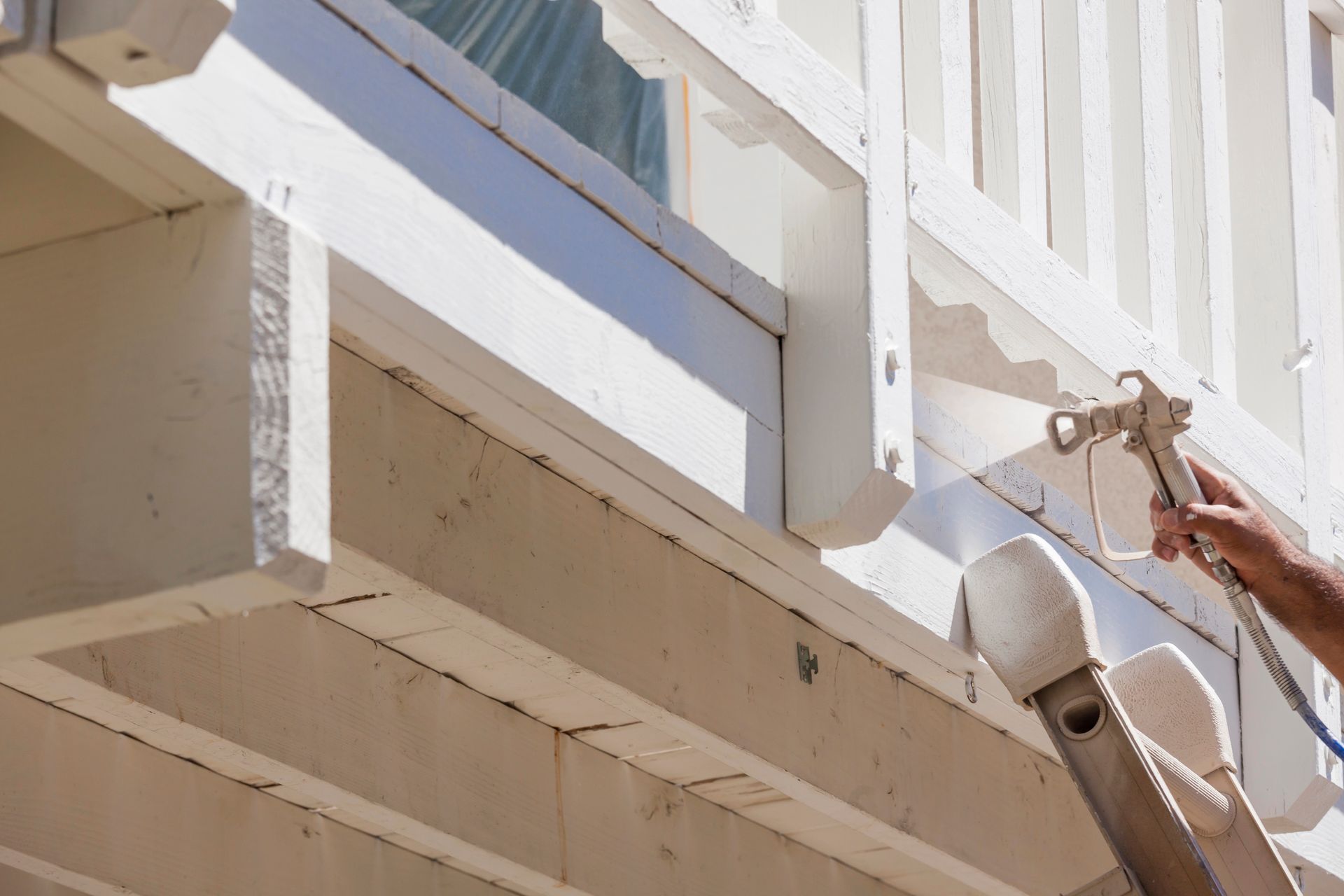 Person spray-painting white paint onto a wooden balcony railing.