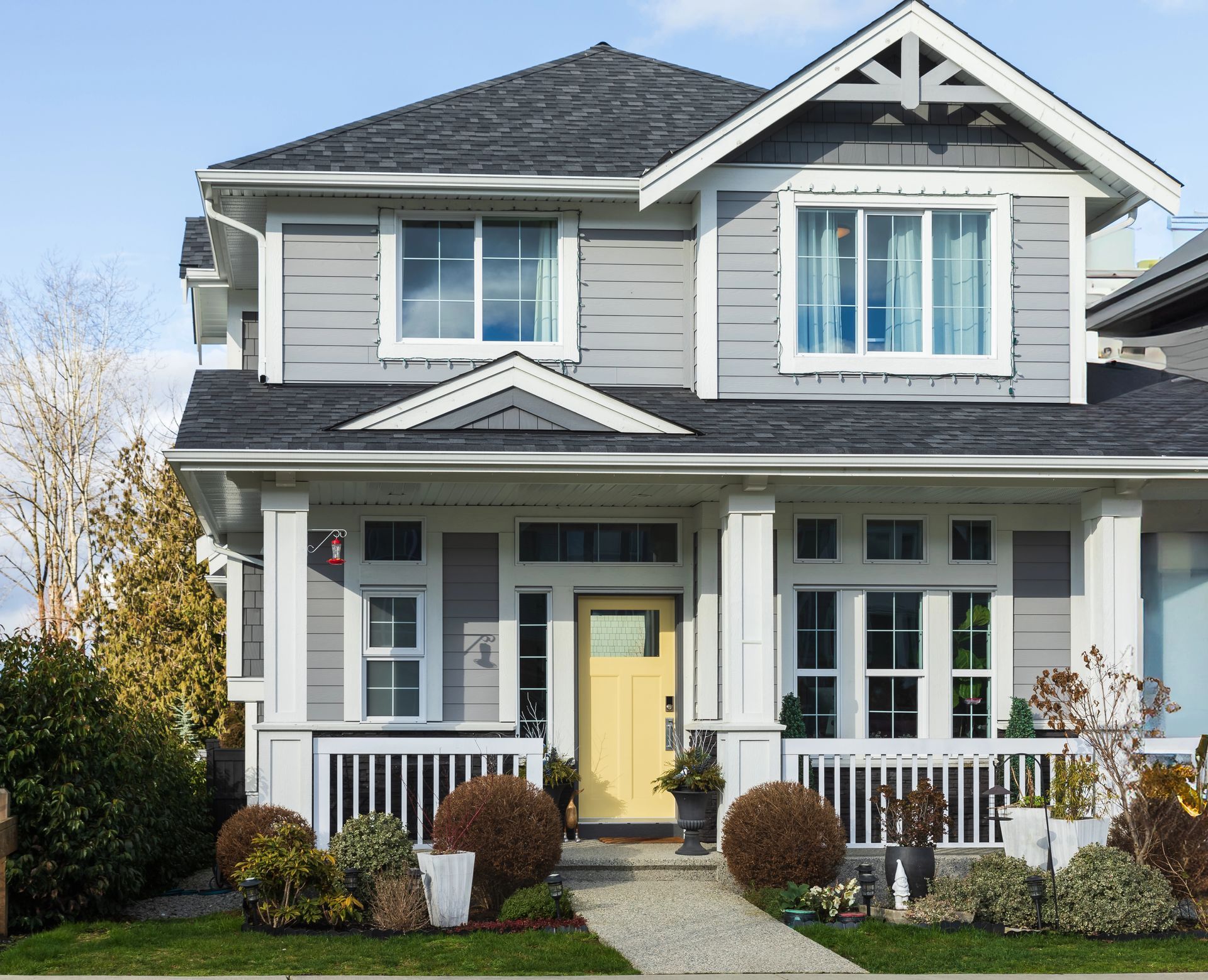 Gray two-story house with white trim, porch, and a yellow front door. Landscaping and a blue sky are present.