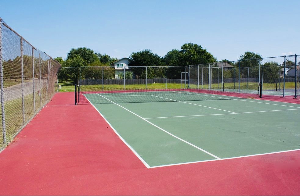 Tennis court with green playing surface, red surrounding area, chain link fence, and blue sky.