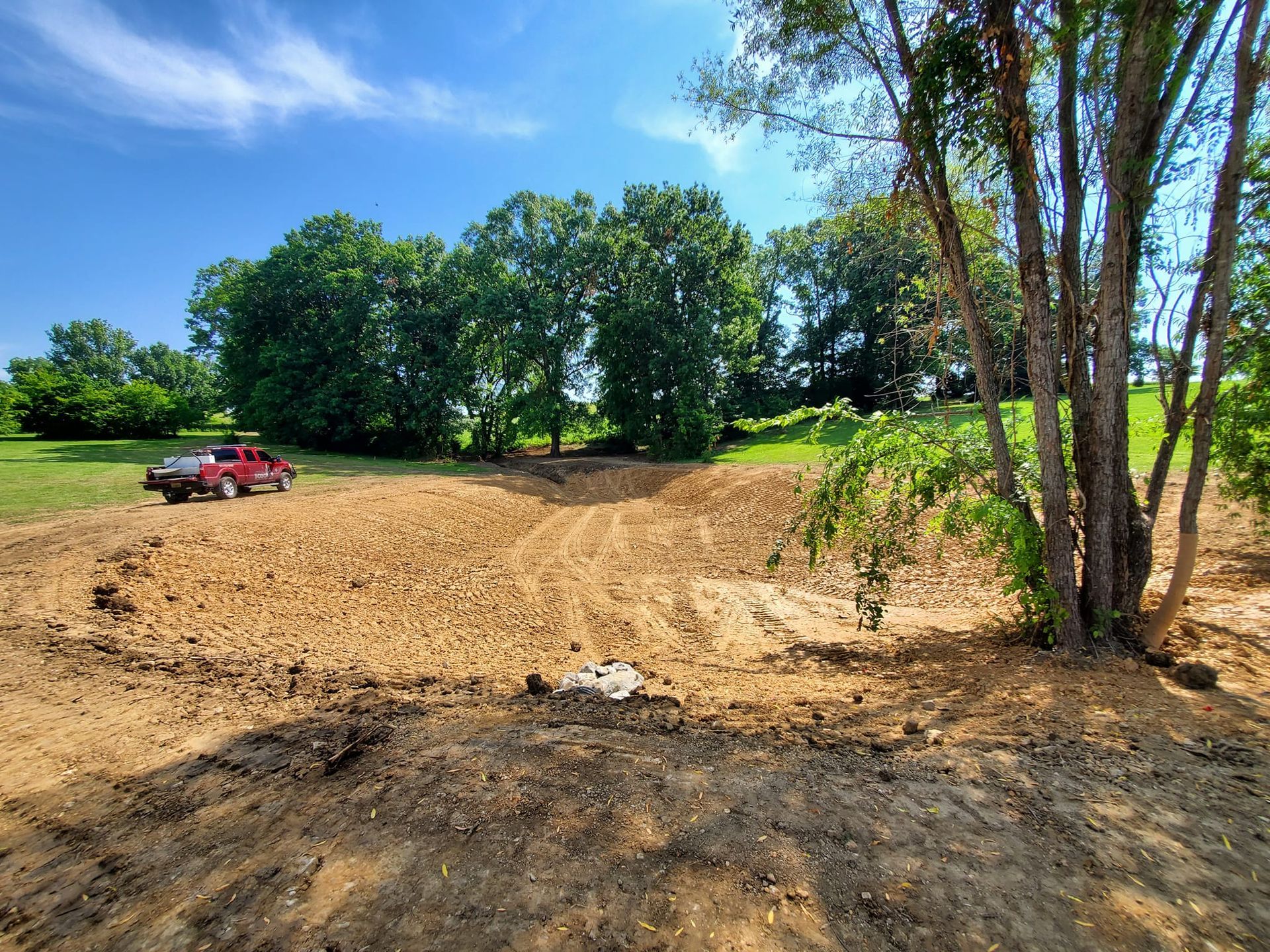 A red truck is parked in the middle of a dirt field surrounded by trees.
