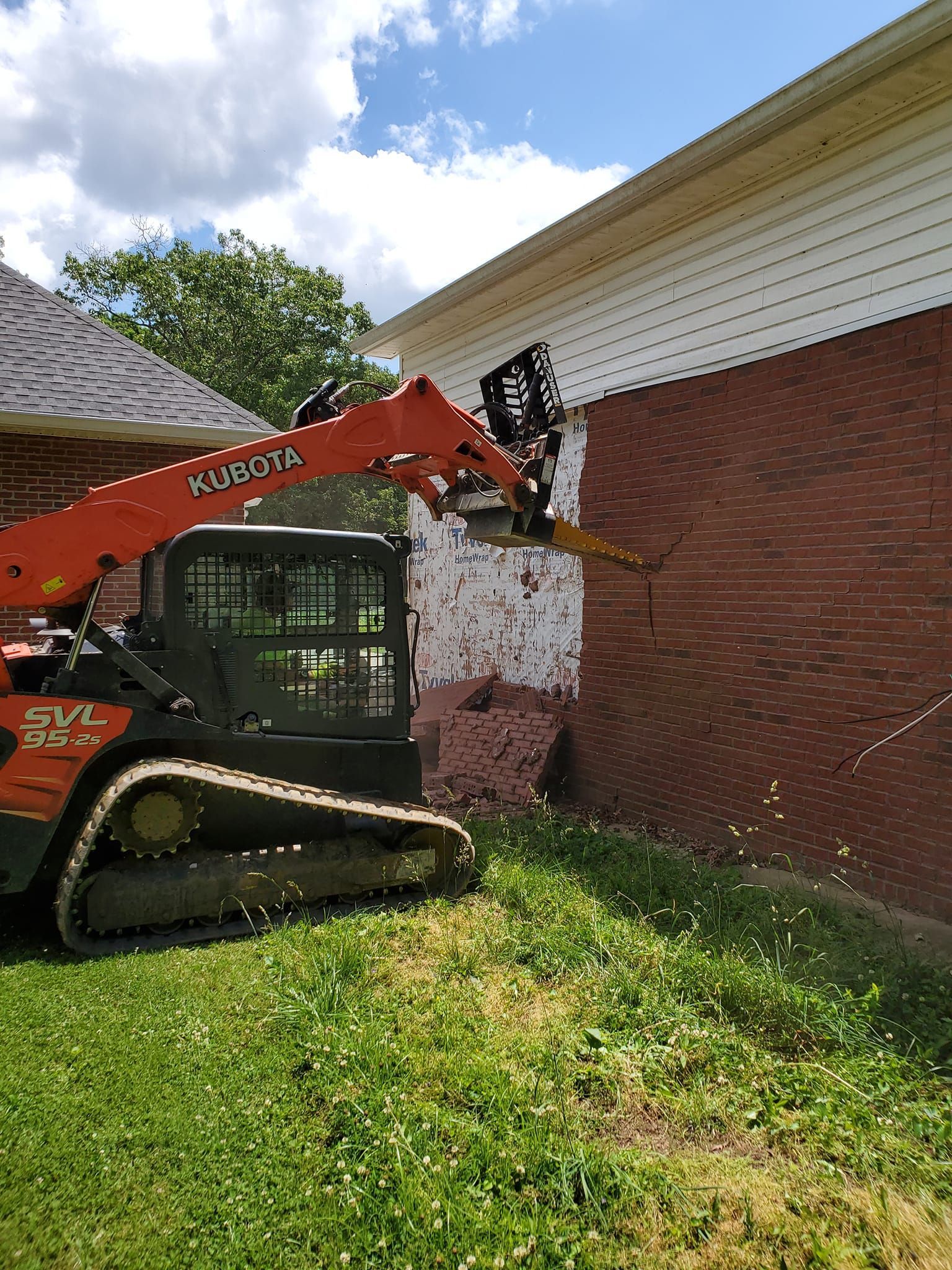 A bulldozer is cutting down a brick wall in front of a house.