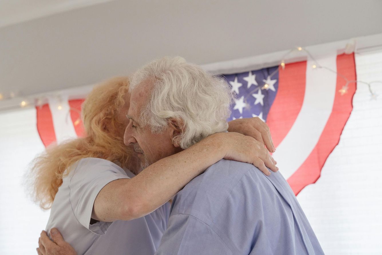An elderly couple is hugging in front of an american flag.