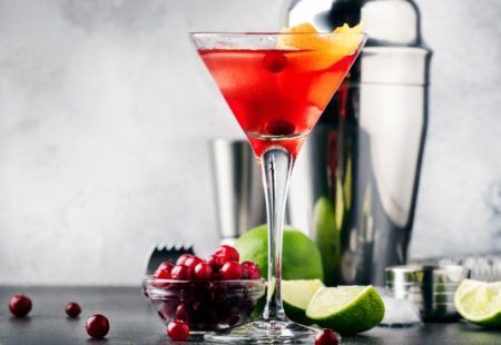 A martini in a martini glass with cranberries and limes on a table.