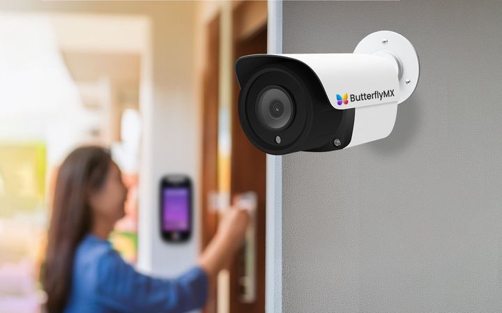 A white security camera mounted on a gray wall overlooking a person interacting with an electronic door access panel.