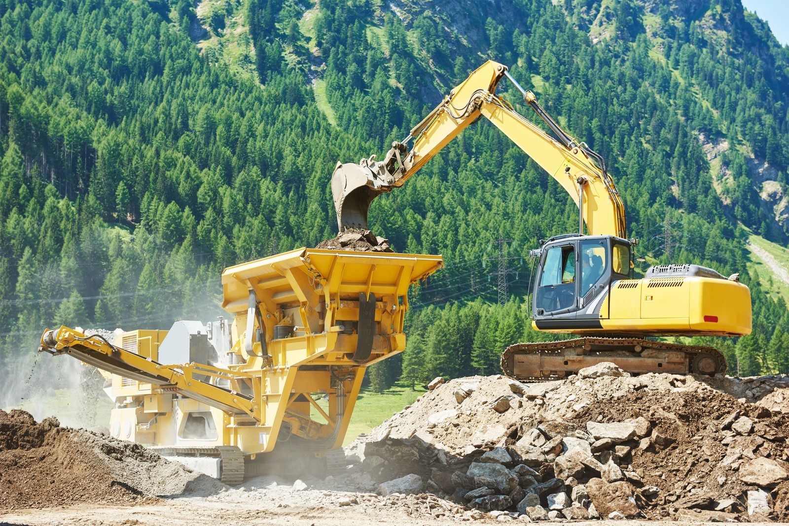 A large yellow excavator is working on a construction site in the mountains.