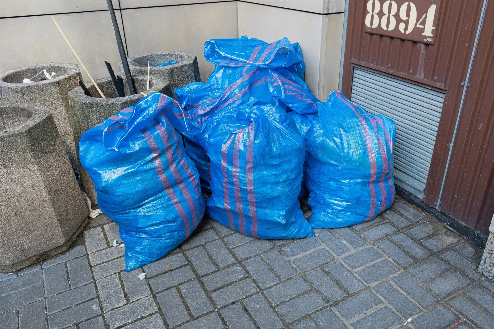 A bunch of blue bags are sitting on a brick sidewalk in front of a building.