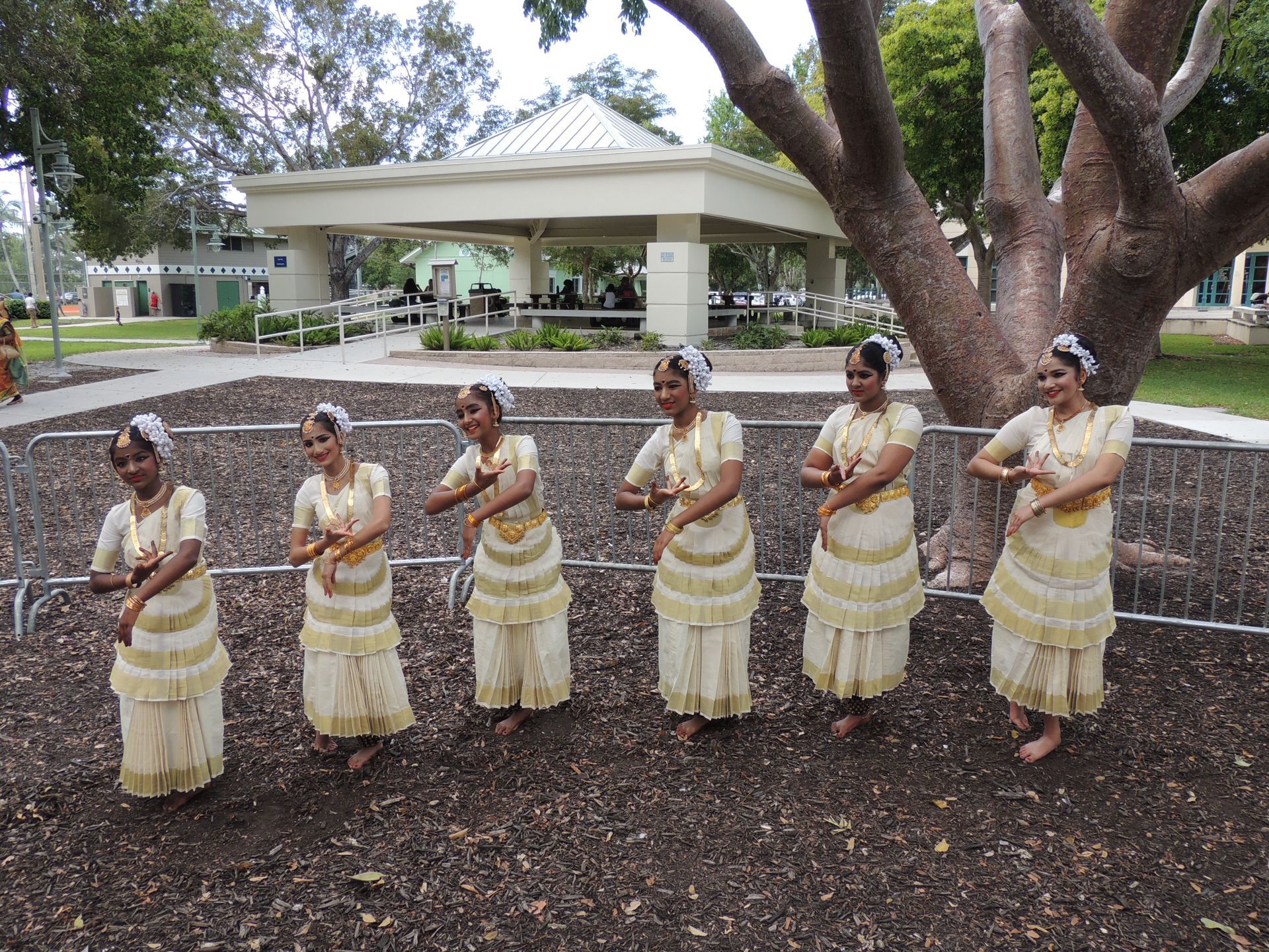 Six women in cream dresses perform a dance outdoors; a gazebo and trees are in the background.