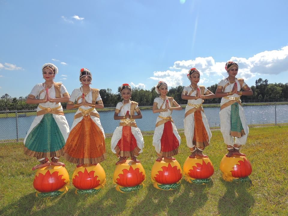 Six dancers in colorful costumes, standing on pots, hands in prayer pose outdoors.