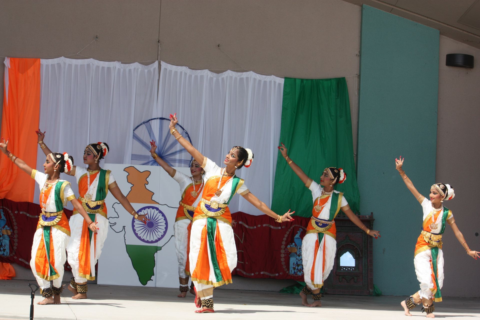 Indian dancers in orange and white costumes perform on stage, in front of the Indian flag.