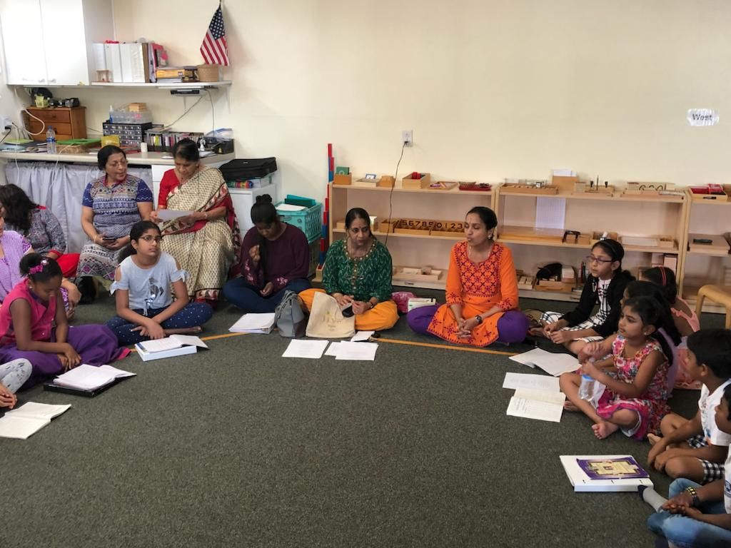 Group of people, adults and children, sit in a circle on a green carpeted floor, papers in front of them.