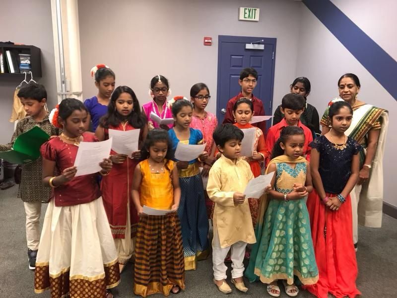 Children in traditional Indian attire singing, holding papers indoors.