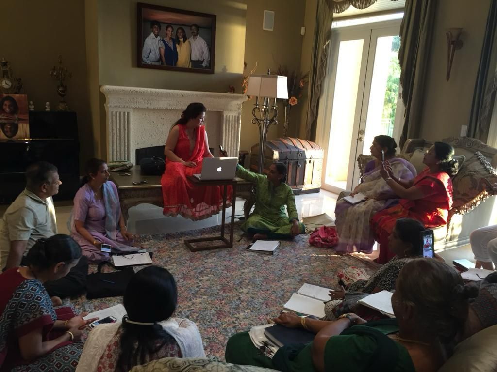 Group of people in colorful attire, circle on the floor, listening to woman presenting with laptop in a living room.