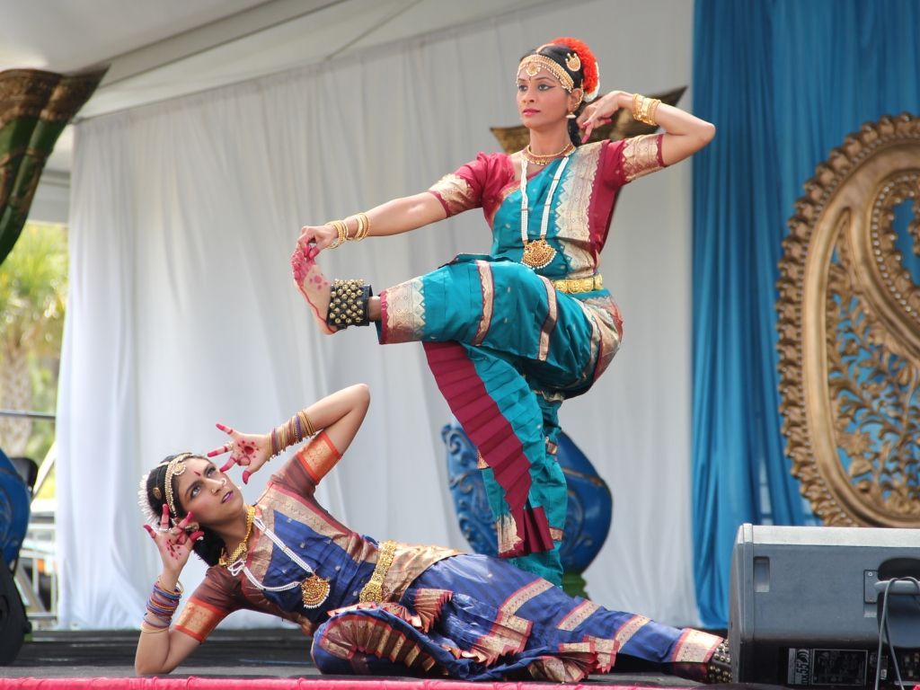 Two women in traditional Indian dance attire perform on a stage. One is standing, lifting a leg. The other is kneeling.