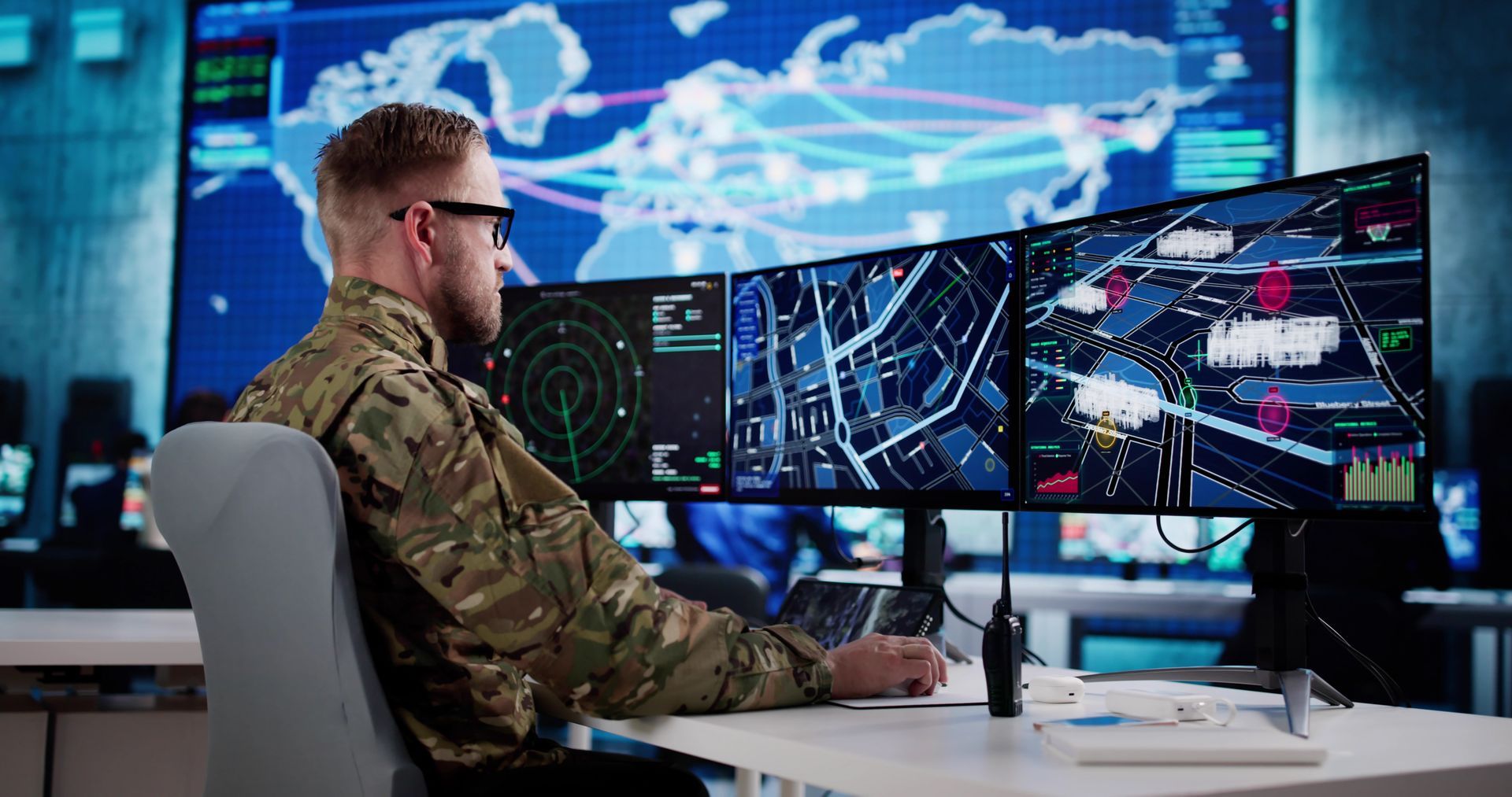 A service member in camouflage uniform monitors digital maps and radar data on three computer screens in a control room.