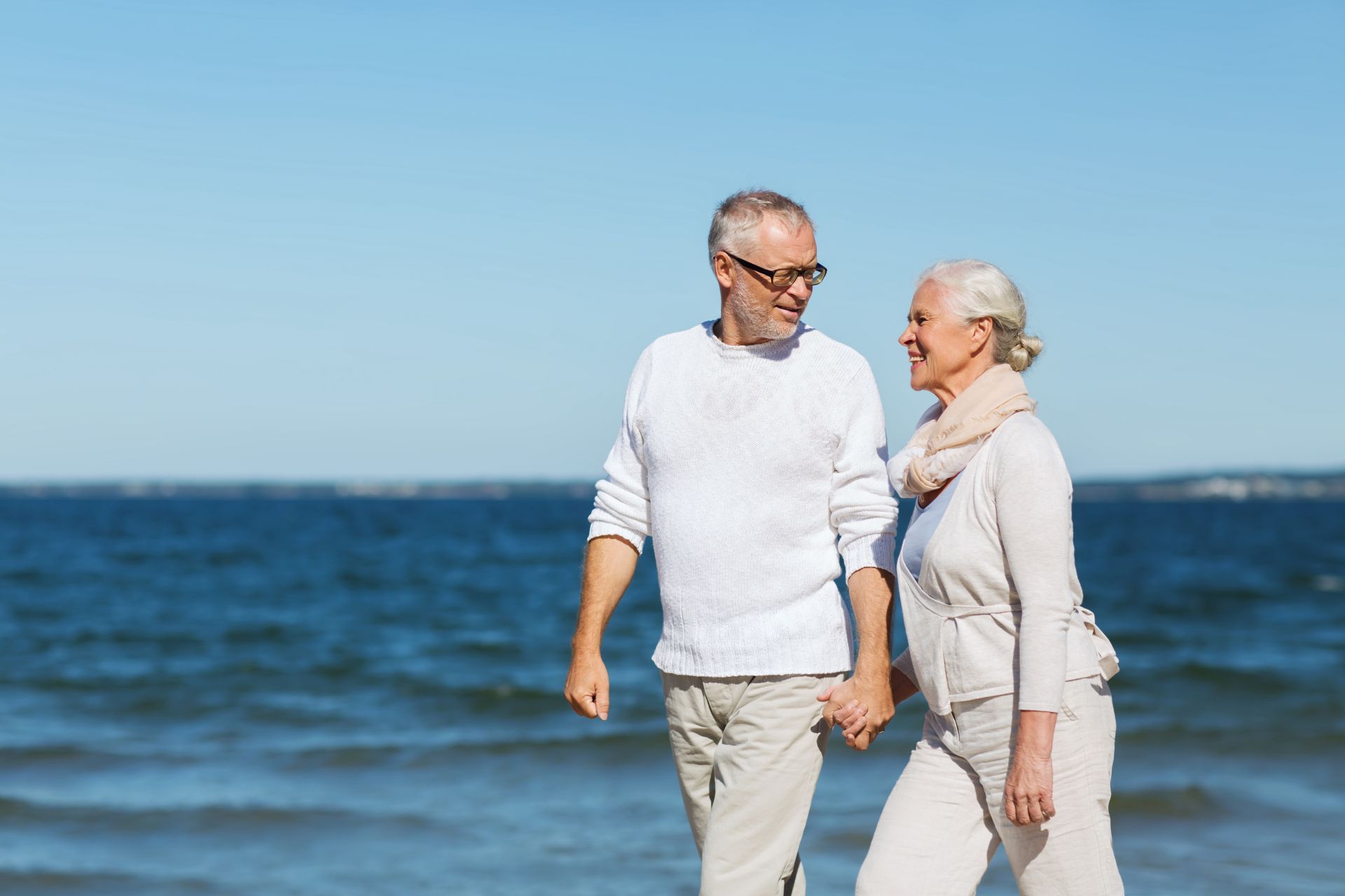 Couple holding hands, walking on a beach, smiling at each other, with blue ocean and sky in background.